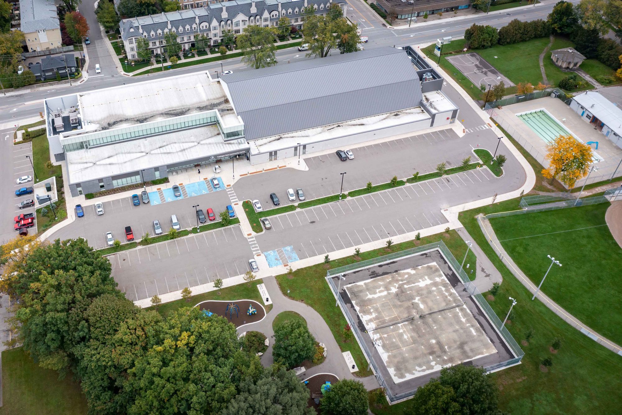 Aerial view of community centre and parking lot with tennis court and playground in the frame.