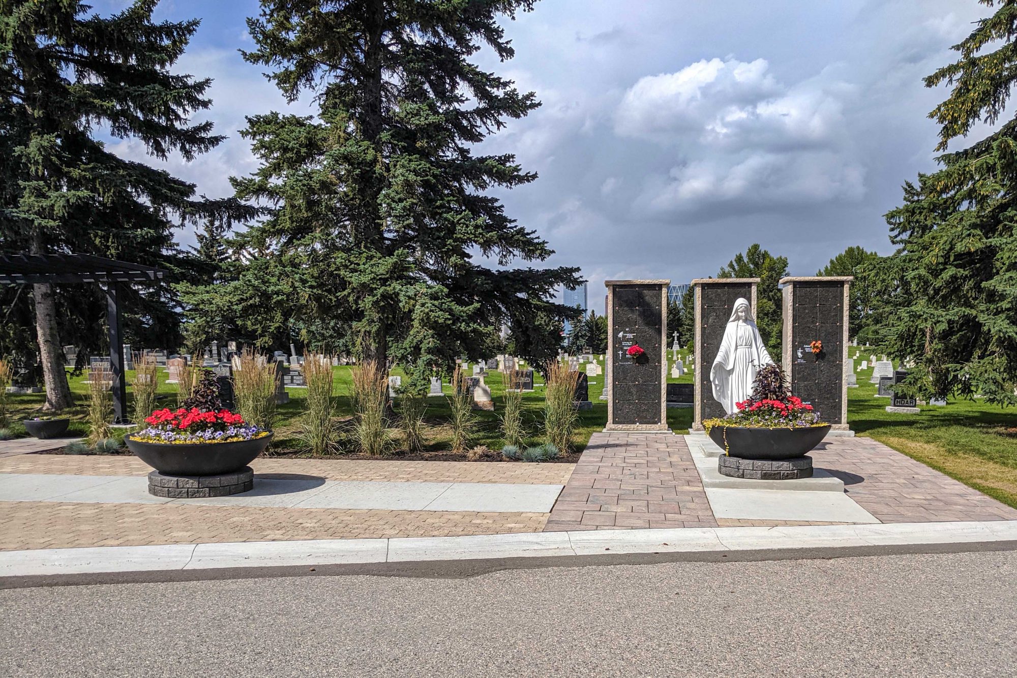 Statue of Mary in the foreground at the entry plaza to a cemetery featuring interlocked stone paving along with plants, flowers and trees. The cemetery and the City of Calgary are in view behind the statue of Mary and the columbaria.