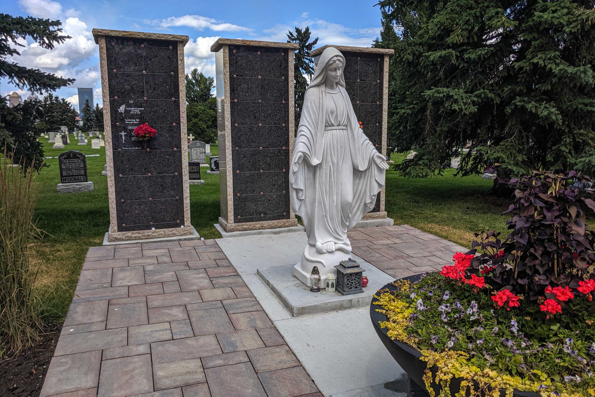 Granite statue of Mary sits in front of columbaria surrounded by trees and plantings. The cemetery is in view behind the columbaria.