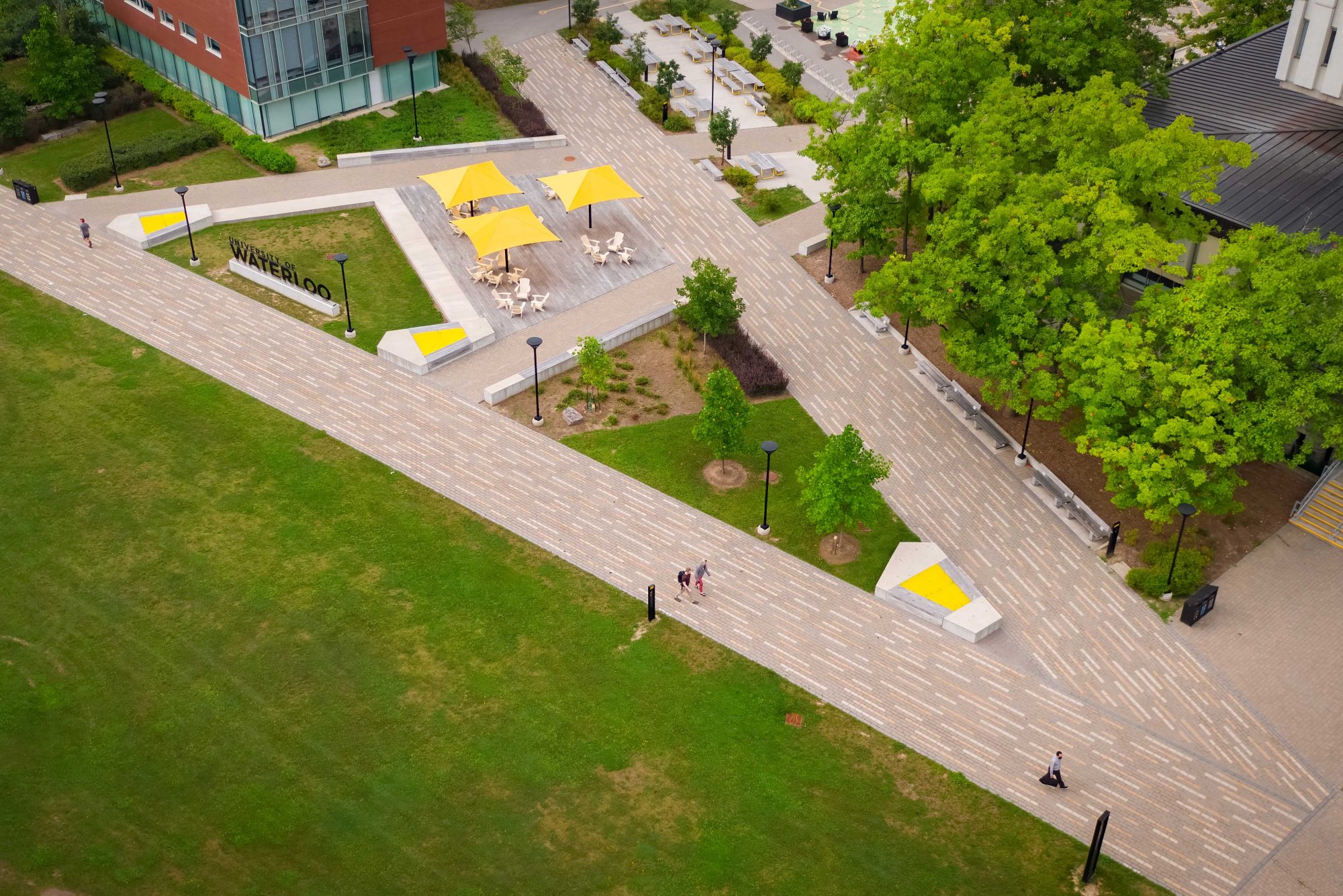 Outdoor common area at the University of Waterloo with three yellow umbrellas and Muskoka chairs. Linear interlocked paving is in the shape of a letter A.