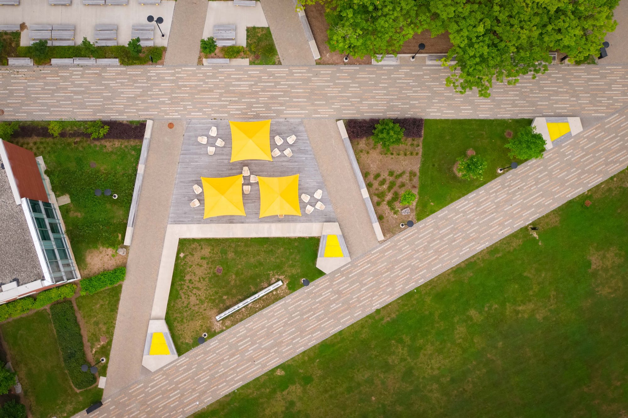 Outdoor common area at the University of Waterloo with three yellow umbrellas and Muskoka chairs. Linear interlocked paving is in the shape of a letter A.