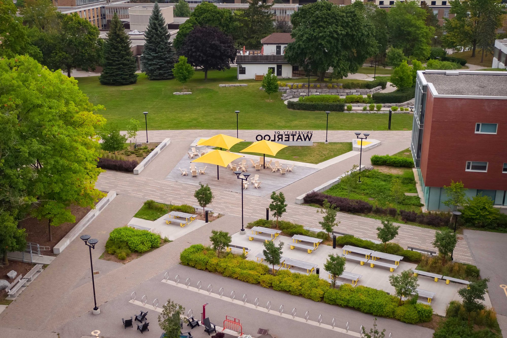 View of the Quad and South Common areas at the University of Waterloo, with three yellow shade umbrellas, Muskoka chairs and wood picnic tables. Bicycle racks are in the foreground.