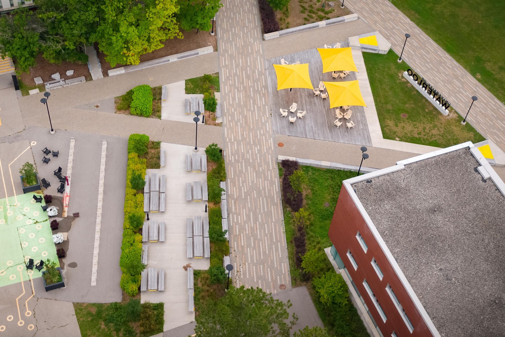 Aerial view of the Quad and South Common areas at the University of Waterloo, with three yellow shade umbrellas, Muskoka chairs and wood picnic tables.