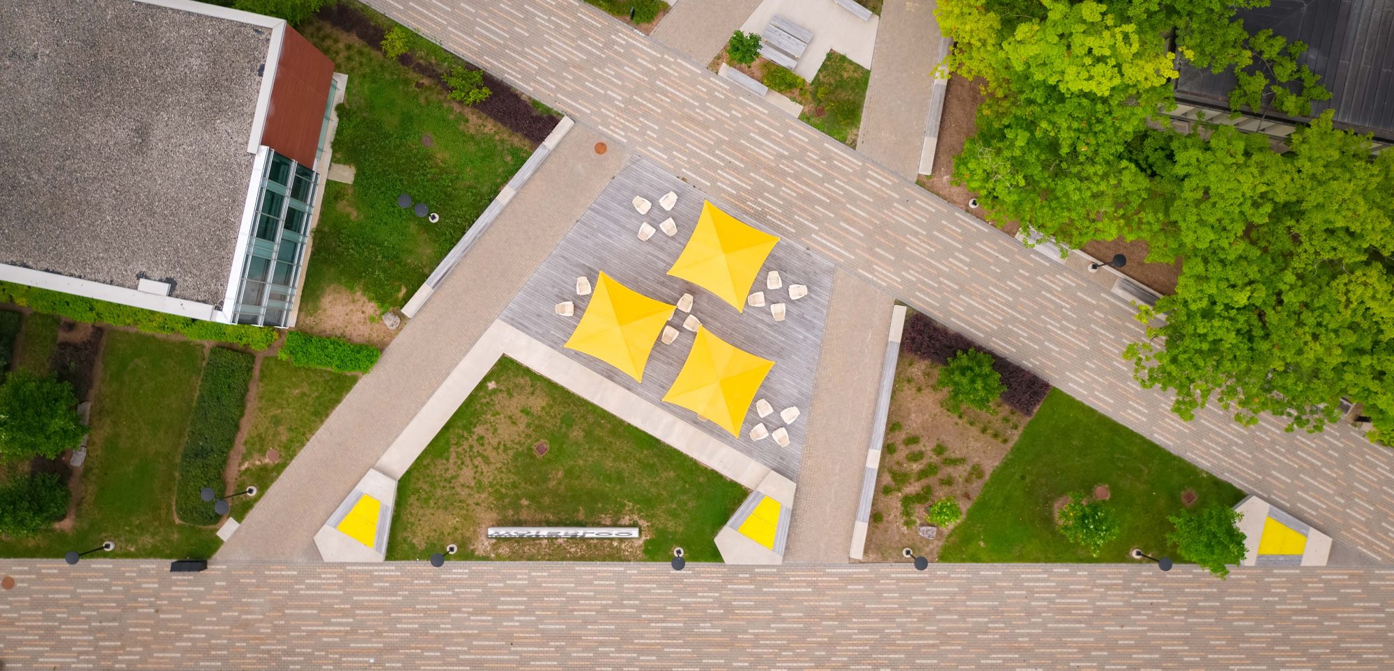 Aerial view of the University of Waterloo South Common grounds with a stones path in the shape of a letter A. Central to the image are three yellow umbrellas with seating.